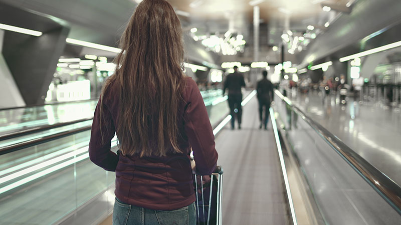 Woman with Luggage Stand on Speedwalk at Airport. Back View of Caucasian Girl with Trolley. Female Tourist on Moving Travelator. People Walking by. Modern Depature Terminal.