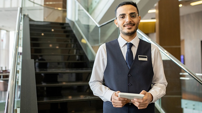 Happy young elegant worker of contemporary hotel using tablet while standing against staircase and looking at you inside lounge